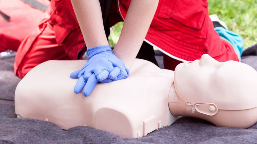 Person in red clothing and blue gloves performing CPR on a practice dummy outdoors. The dummy is lying on a dark surface, and the person is applying chest compressions with interlocked hands.