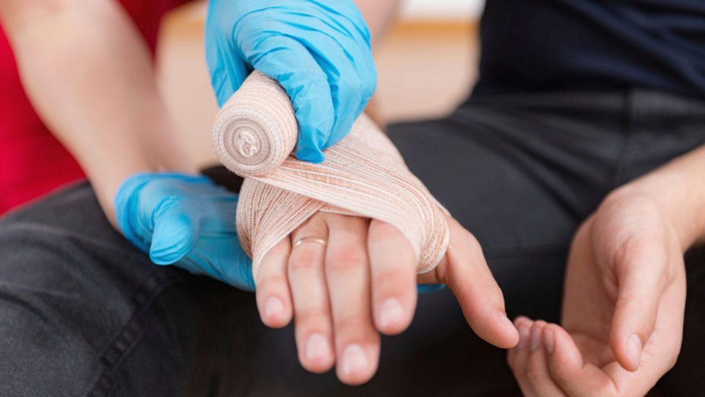 A person with gloved hands is wrapping a white bandage around another person's injured hand. The seated person has their hands resting on their lap, and the background is out of focus.