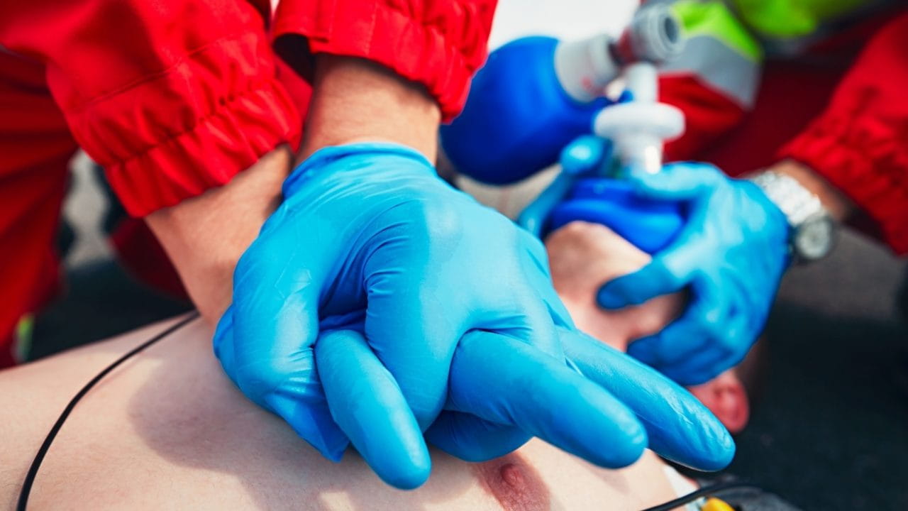 A person wearing blue gloves performs CPR chest compressions on another individual lying on the ground. A second person assists with a bag-valve mask in the background, providing ventilation. Both wear red uniforms, indicating emergency medical services.