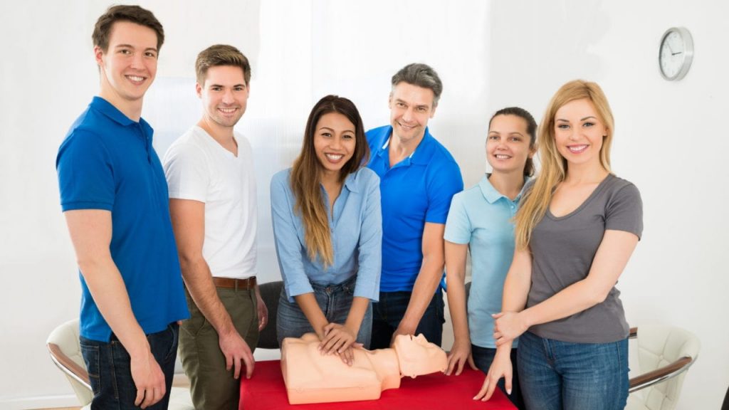 A group of six people smiles near a CPR mannequin on a table. One person demonstrates CPR by pressing on the mannequin's chest. They are in a bright room, with casual attire. A clock is visible on the wall.
