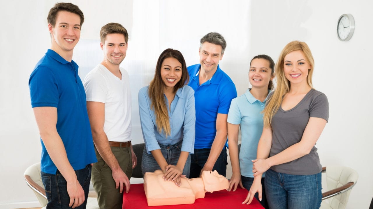 A group of six people smiles near a CPR mannequin on a table. One person demonstrates CPR by pressing on the mannequin's chest. They are in a bright room, with casual attire. A clock is visible on the wall.