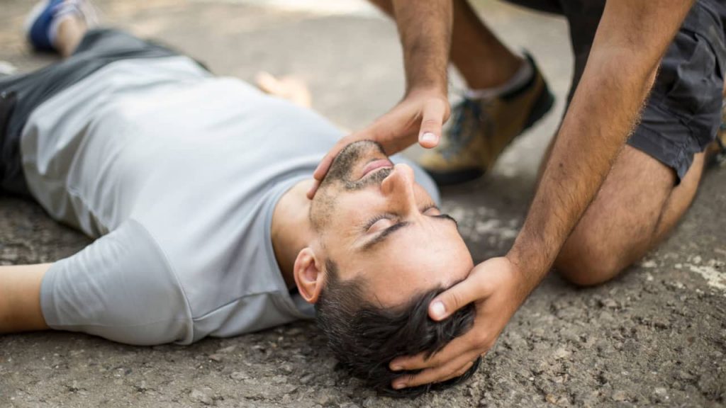 A person lies on the ground with their eyes closed, wearing a light gray t-shirt. Another individual, kneeling beside them, supports the head and neck with both hands in what appears to be a first aid or emergency assistance scenario.