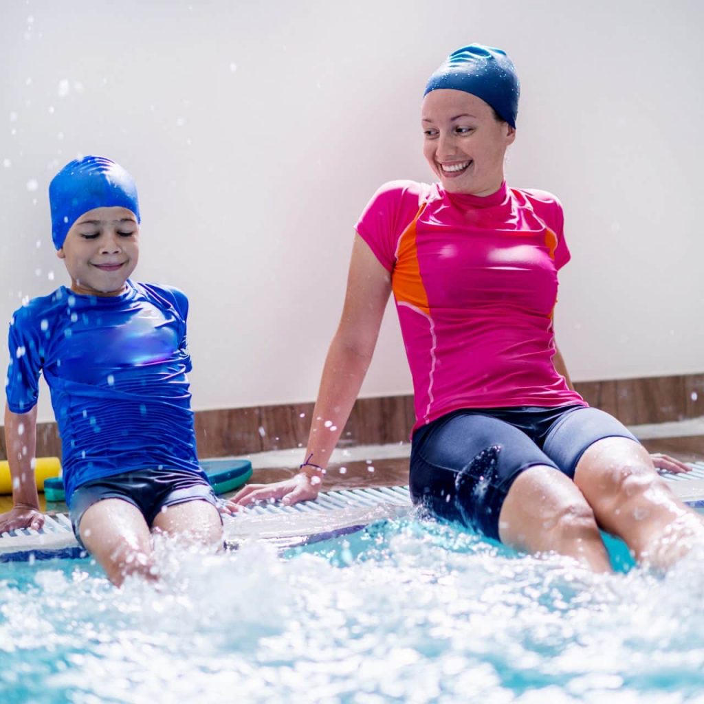 A child and an adult sit at the edge of a pool, splashing water. The child wears a blue swim cap and outfit, while the adult wears a pink and orange top with black shorts. Both are smiling and enjoying the water.