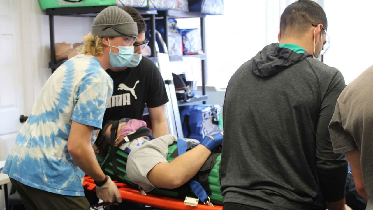 A group of people, wearing masks and gloves, are helping a person on a stretcher. The person on the stretcher is secured with straps, and the setting appears to be indoors, possibly in a medical or training facility.
