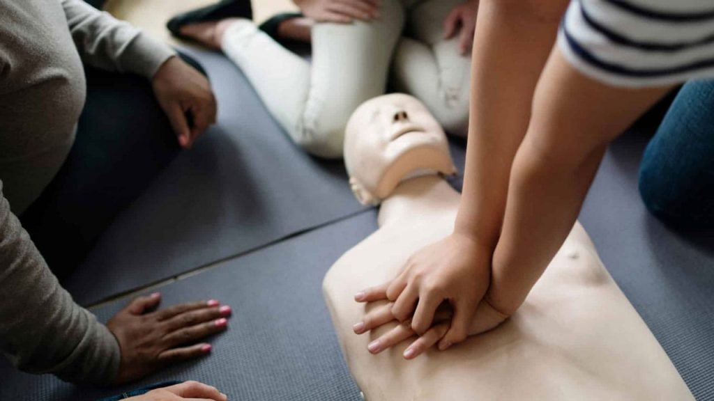 People kneeling on the floor practice CPR on a manikin. One person's hands are performing chest compressions. Others watch attentively, demonstrating a CPR training session.