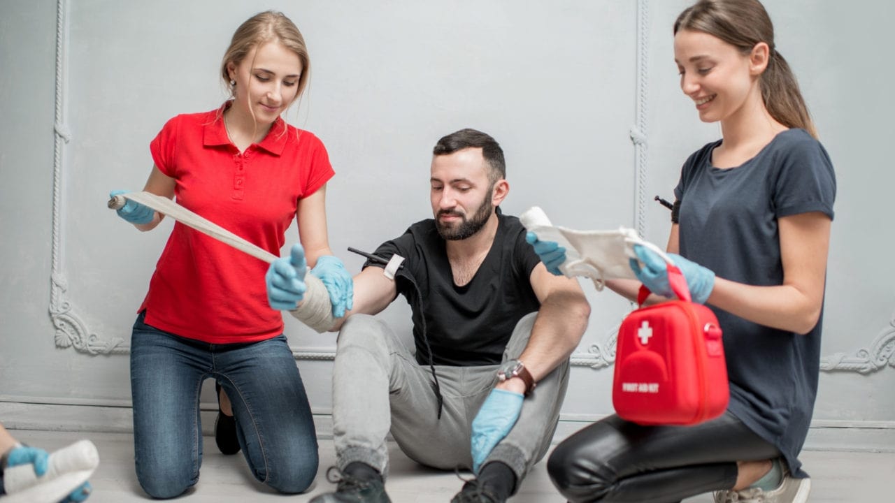 Two women in casual attire and gloves assist a seated man with a bandaged arm. One woman is holding the bandage, and the other has a first aid kit. They are indoors against a light-colored wall.