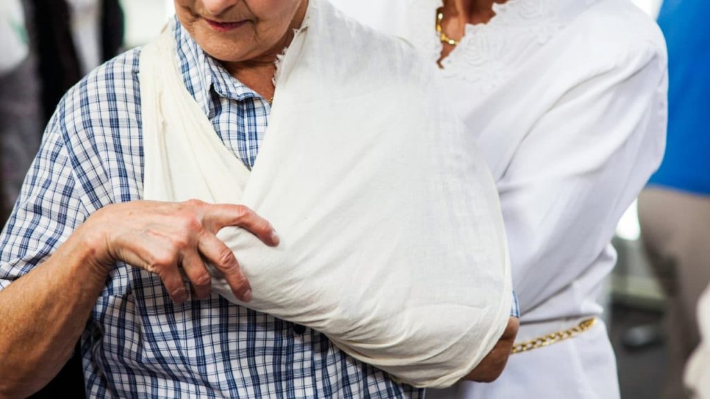 An elderly person wearing a blue checkered shirt has their arm in a white sling. A person in white clothing is assisting them from behind, holding the sling gently. The background is blurred, with hints of other people present.