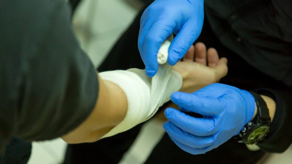 A person wearing blue gloves is wrapping a white bandage around another person's wrist, providing first aid. The scene is focused on the hands and bandaging process.