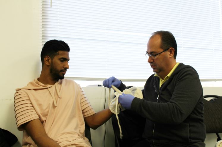 A man wearing a pinkish beige shirt is seated as another man, wearing glasses and blue gloves, wraps a bandage around his arm. They are indoors, in front of a window with blinds, and appear focused on the task at hand.