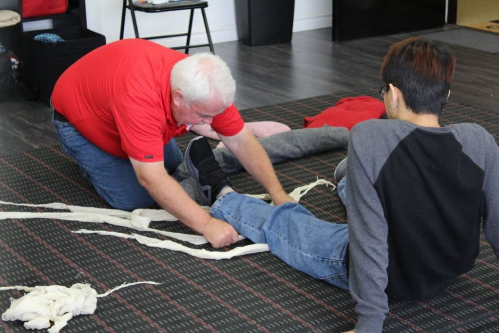 An older man in a red shirt kneels on the floor while demonstrating how to wrap a bandage around a younger person's leg. The younger person, wearing a gray and black shirt, sits on the floor with their leg extended. Medical training supplies are scattered around them.