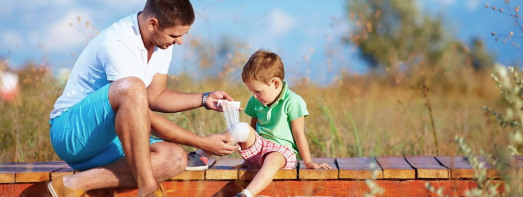 A man is assisting a child by wrapping a bandage around the child's knee. They are sitting on a wooden bench in a sunny outdoor setting, surrounded by grass and trees. The man is wearing a white shirt and blue shorts, and the child is in a green shirt.