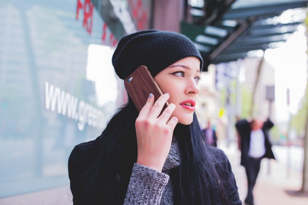 A woman with long dark hair, wearing a black beanie and coat, holds a smartphone to her ear while standing on a city sidewalk. She looks to the side, with blurry buildings and pedestrians in the background.