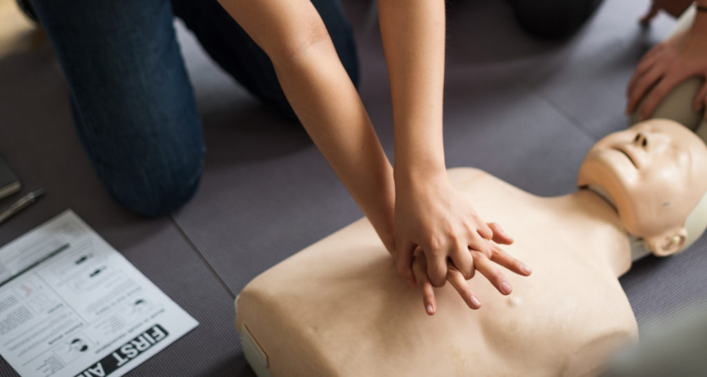 Person practicing CPR on a mannequin, pressing down on the chest with interlocked hands. A first aid manual is visible on the side, and the setting appears to be an indoor training session.