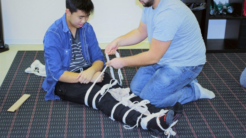 A man using medical gauze to wrap another man’s injury, assisting him in standing up during a first aid training session.