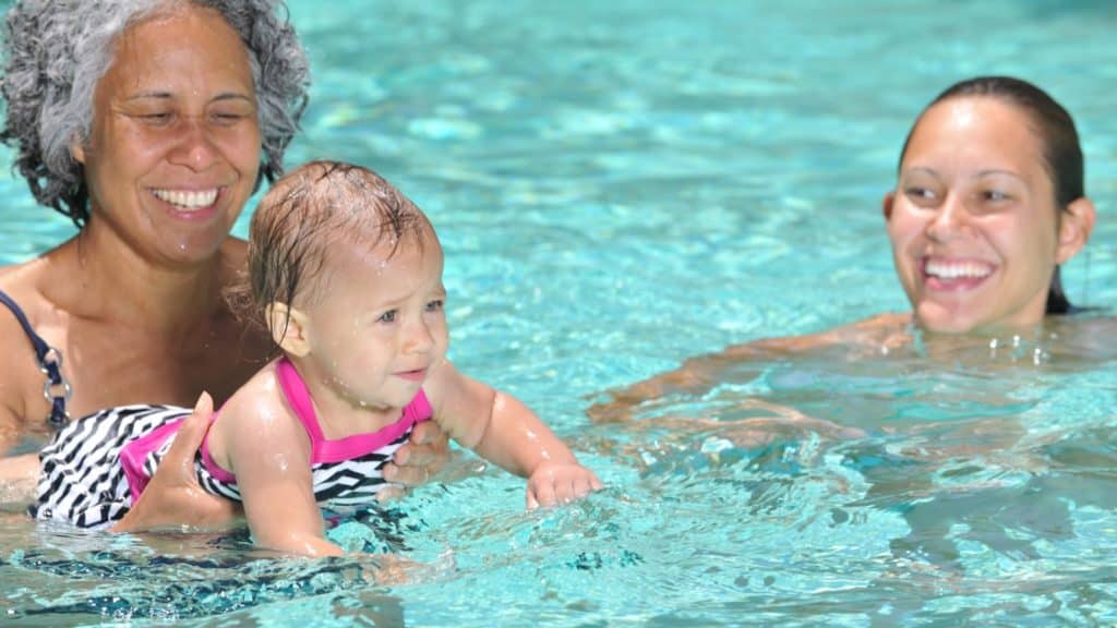 A baby in a pink swimsuit is held up in a swimming pool by an older woman with gray hair and a black-and-white striped swimsuit. A younger woman with brown hair smiles in the water next to them, all enjoying the sunny day.