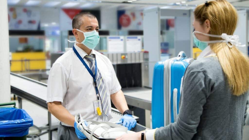 A security officer wearing a mask and gloves speaks with a traveler, also wearing a mask, at an airport security checkpoint. The traveler is holding a suitcase and a laptop tray. Conveyer belts and security equipment are visible in the background.