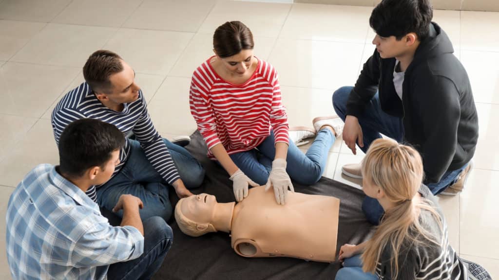A group of five people sitting on the floor in a circle, practicing CPR on a mannequin. The instructor wears gloves and demonstrates chest compressions. The participants watch attentively. They are on a tiled floor with casual attire.