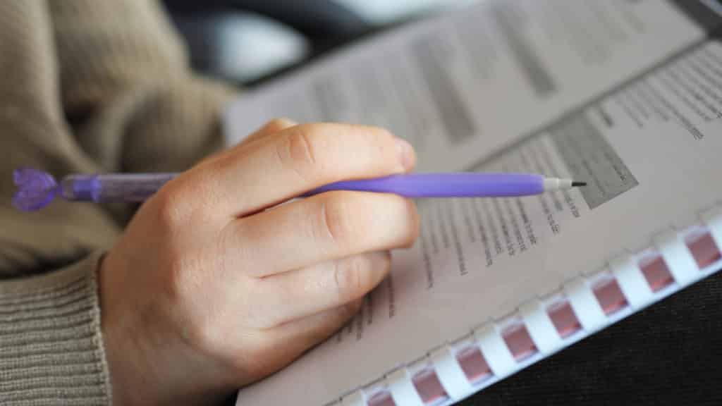 A person holds a purple pen, poised to write on an open document. The paper has text and some highlighted sections. The person's sleeve is beige, suggesting casual attire.
