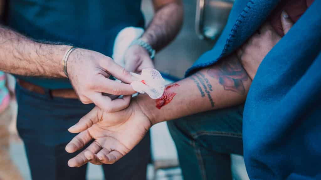 A person is treating a bleeding wound on another person's arm using a piece of white gauze. The injured person wears a blue fabric and has visible tattoos on their forearm. The focus is on the care process.