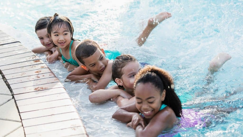 Five children smiling and playing at the edge of a swimming pool, with water splashing around them. They are lying on their stomachs in the shallow water, showing joy and excitement in a bright, sunny setting.
