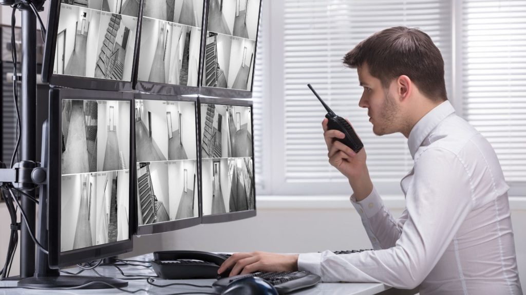A man in a white shirt is seated at a desk, monitoring security footage on multiple screens. He holds a walkie-talkie, appearing focused on the surveillance images displayed in front of him.
