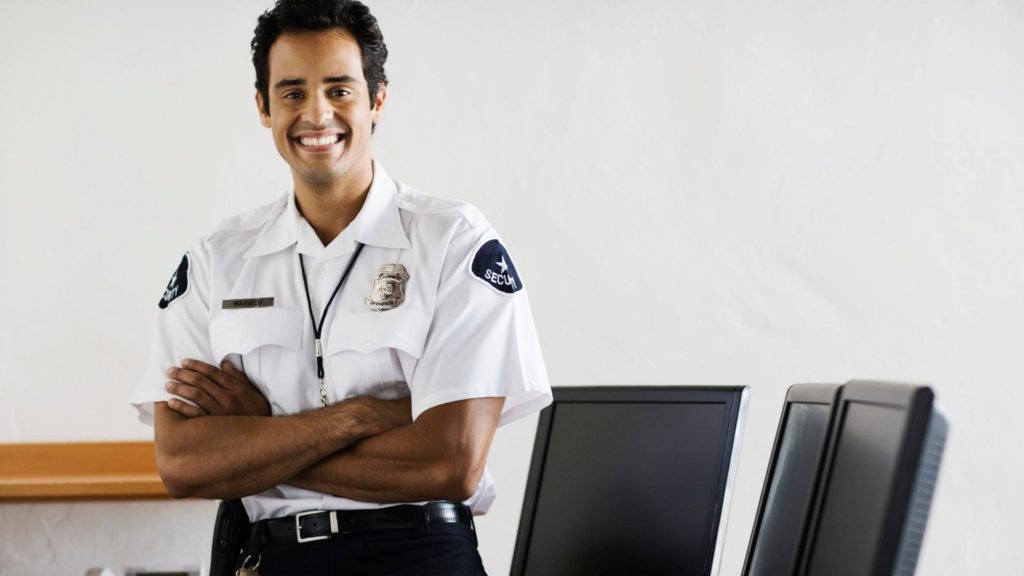 A security guard in a uniform with a badge stands smiling inside an office, arms crossed. Two computer monitors are on a desk beside him. The background is a plain, light-colored wall.