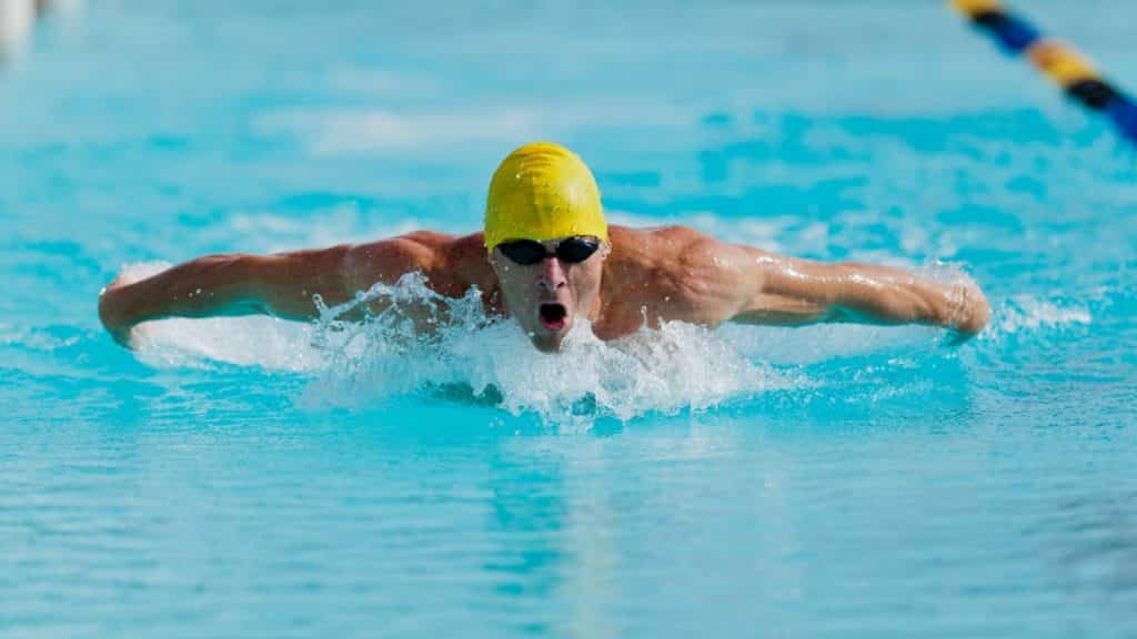 A swimmer wearing a yellow cap and goggles is performing the butterfly stroke in a bright blue swimming pool. Water splashes around as they advance, showcasing dynamic motion and focus.