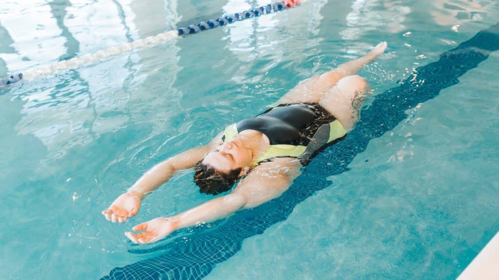A person in a black and yellow swimsuit is swimming on their back in a clear indoor pool, with arms extended overhead. The water is calm, and lane markers are visible in the background.