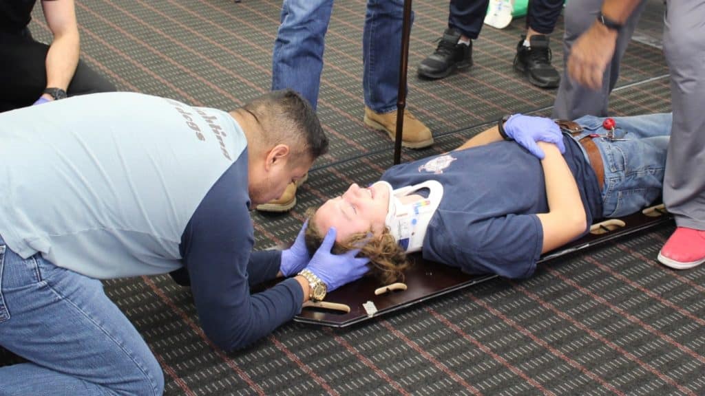 A person wearing a neck brace is lying on a backboard while another person kneels beside them, holding their head. Several people are standing nearby, all wearing gloves, on a carpeted floor.