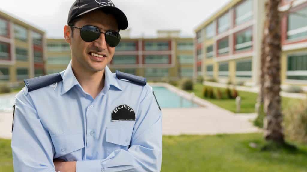 A security guard in a light blue uniform and sunglasses stands with arms crossed. He is smiling. In the background, there are modern buildings, a pool, and a grassy lawn with a palm tree.