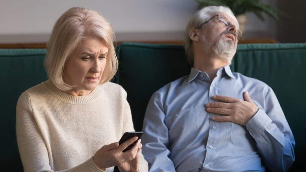 An elderly woman looks at her smartphone, appearing concerned. Next to her, an elderly man is leaning back on a couch, eyes closed, with one hand on his chest.