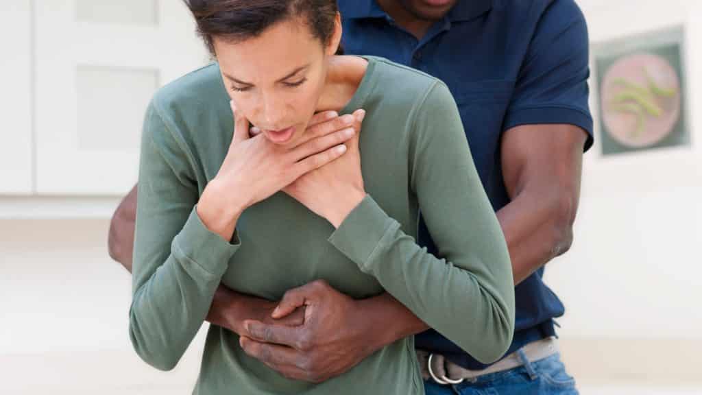 A person in a green shirt appears to be choking, holding their throat. Another person in a blue shirt is performing the Heimlich maneuver, standing behind and applying pressure to their abdomen in a kitchen setting.