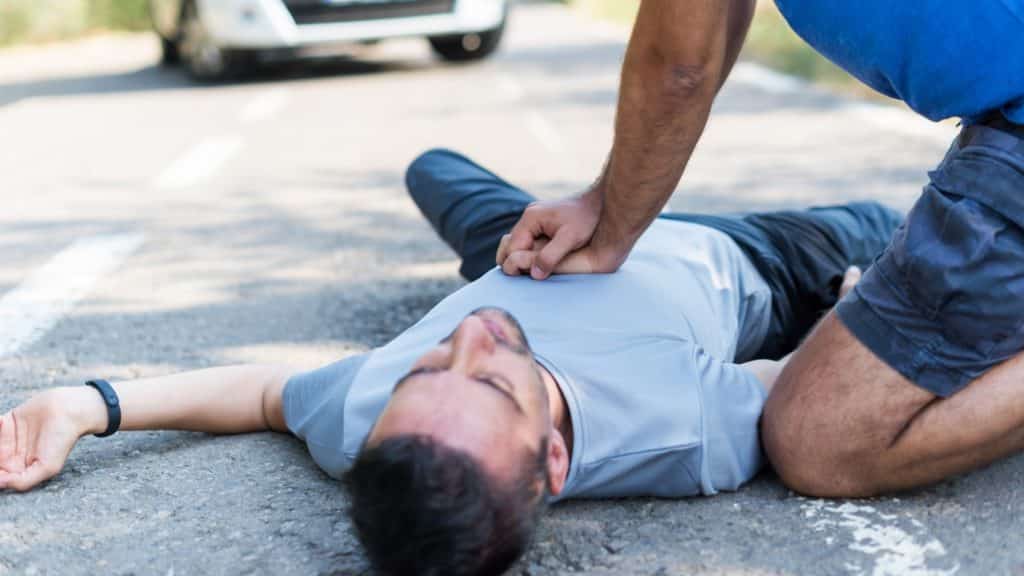 A man lying on a road receives CPR from another person kneeling beside him. The scene suggests a medical emergency. A blurred vehicle is visible in the background.