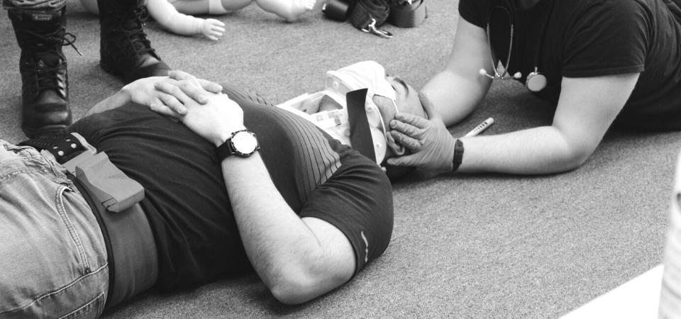 A person lying on the floor with eyes closed, wearing a watch. Another person, wearing a stethoscope, gloves, and a medical mask, is supporting their head and checking their breathing. Medical equipment is visible nearby.