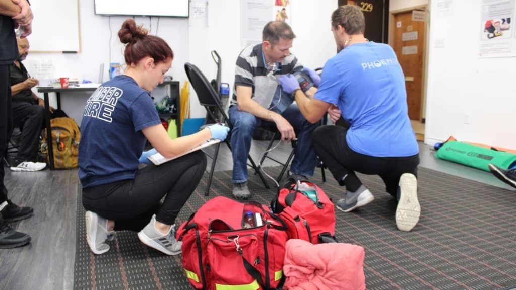Three people in a room during a medical training exercise. Two individuals in blue shirts assist a seated man, wrapping a bandage around his forearm. Medical equipment and bags are on the floor. This appears to be a first aid or emergency scenario practice.