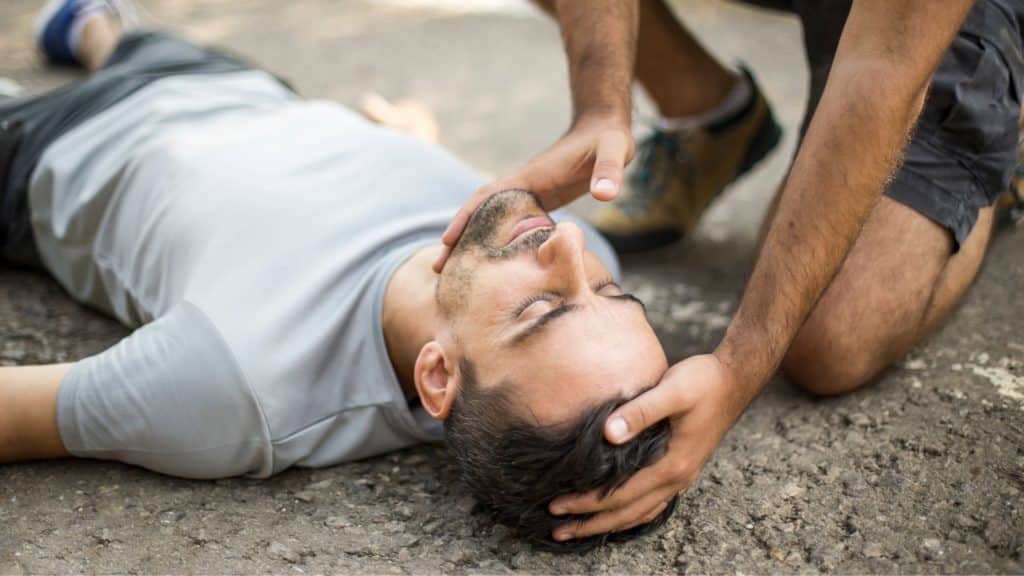 A person lies on their back on a paved surface, eyes closed. Another person kneels nearby, supporting the first person's head with both hands, possibly administering aid.