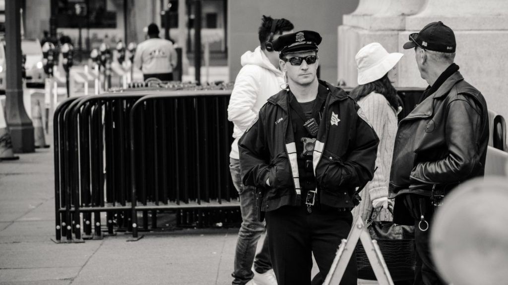 A police officer stands near a barrier on a city sidewalk, wearing a uniform with a cap and sunglasses. People walk by, including a person in a white jacket and another in a hat. The scene is in black and white.