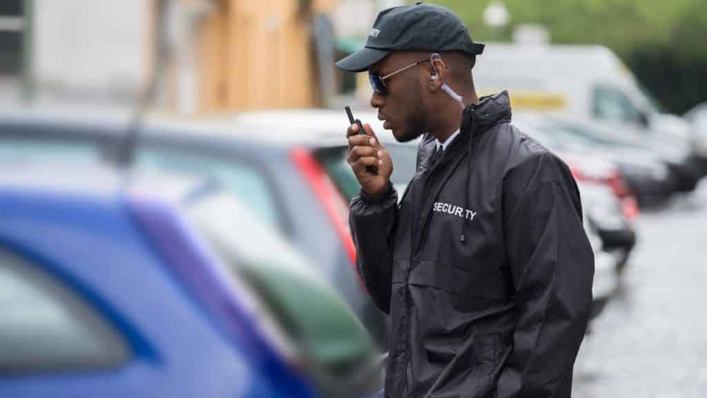 A security guard wearing a black jacket and cap speaks into a walkie-talkie. He stands next to parked cars on a cobblestone street, with buildings in the background.