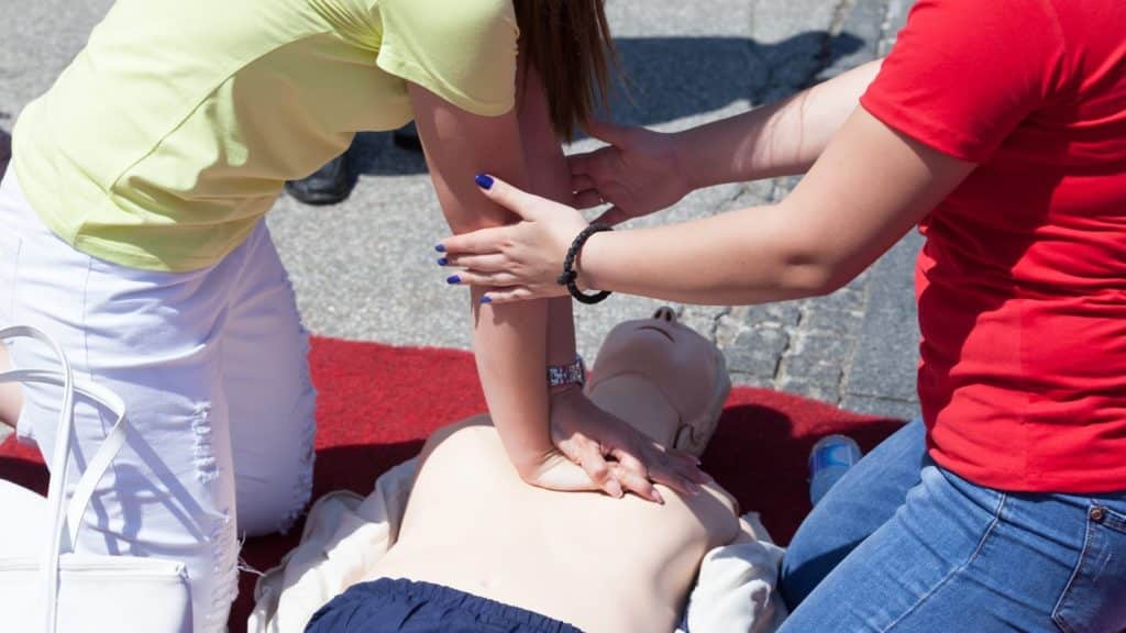 Two people demonstrate CPR on a manikin lying on a red mat outdoors. One person in a yellow shirt performs chest compressions, while another in a red shirt observes closely, adjusting the technique if needed.