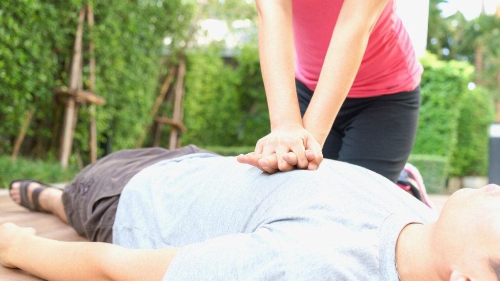 A person in a pink shirt is performing chest compressions on someone lying on their back outdoors. Lush green plants in the background.