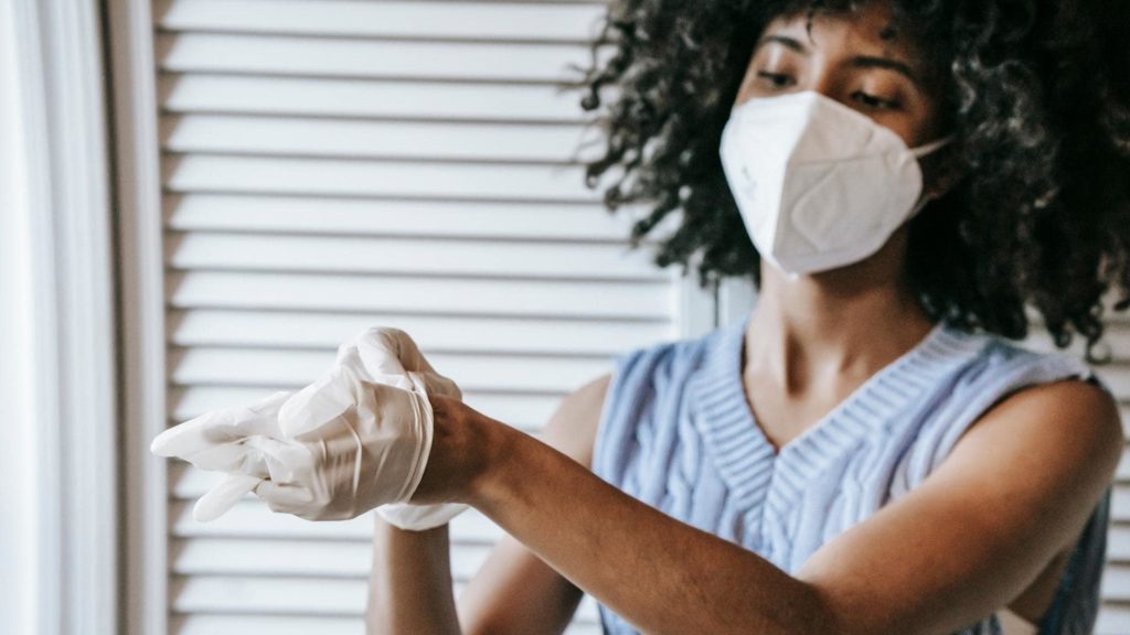 A person wearing a face mask and a light blue sleeveless top is putting on white gloves. They are standing indoors near a window with white blinds.