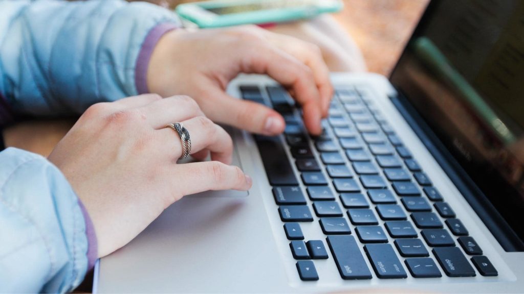A person wearing a bracelet and a ring types on a laptop keyboard. Their hands rest on the keyboard, and a smartphone is partially visible in the background. The person is wearing a light-colored jacket.
