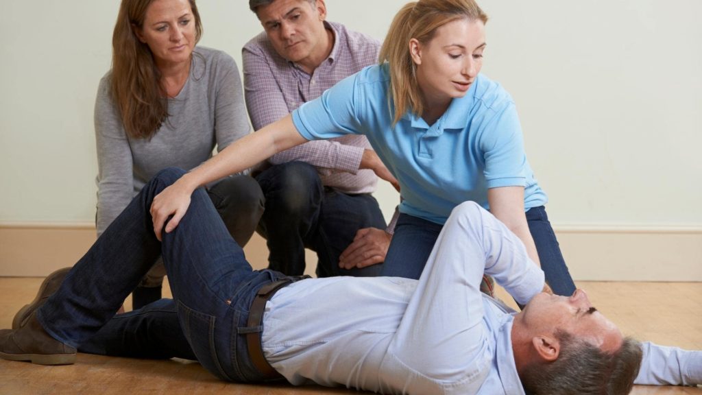 A woman in a blue shirt kneels beside a man lying on the floor, checking his condition. Two other people, a woman and man, watch with concerned expressions. The scene suggests a first aid or emergency situation indoors.