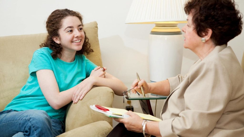 A young woman in a teal shirt sits on a sofa, smiling and talking to an older woman who is holding a pen and notepad. The older woman, seated on another chair, is listening attentively. A lamp is on the table between them.