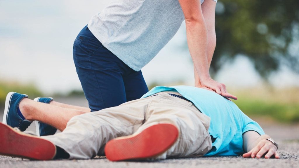 A person in a gray shirt and dark shorts performs CPR on another person lying on the ground, who is wearing a blue shirt and beige pants. The scene takes place outdoors with a blurred background.