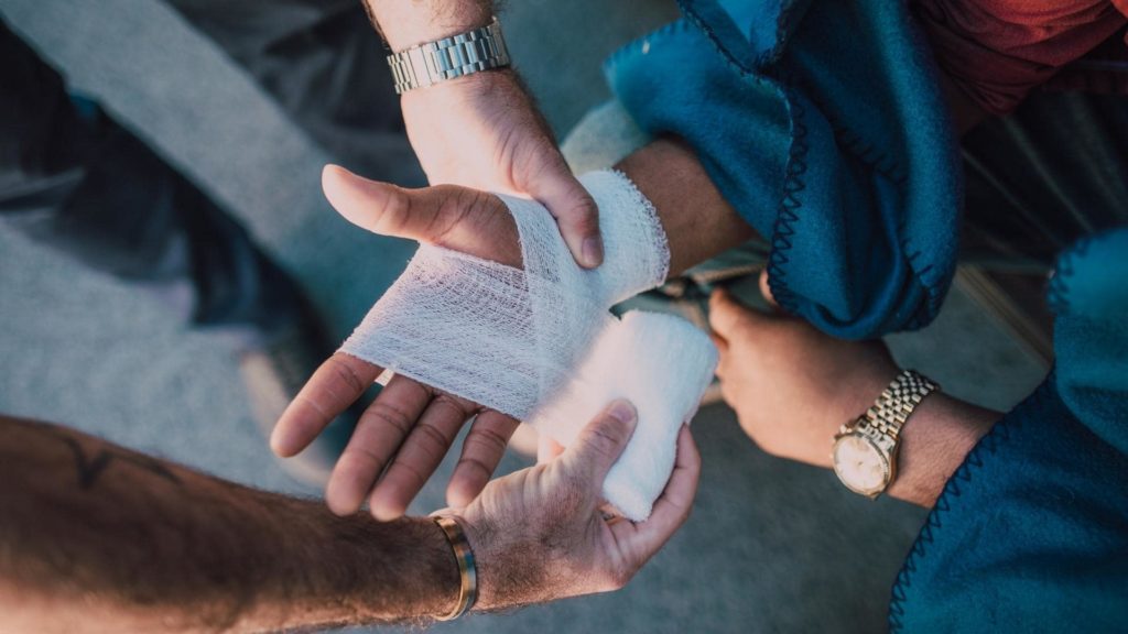 A person is receiving first aid on their hand, which is being wrapped with a bandage by another individual. Both wear watches, and the person receiving care is dressed in blue fabric.