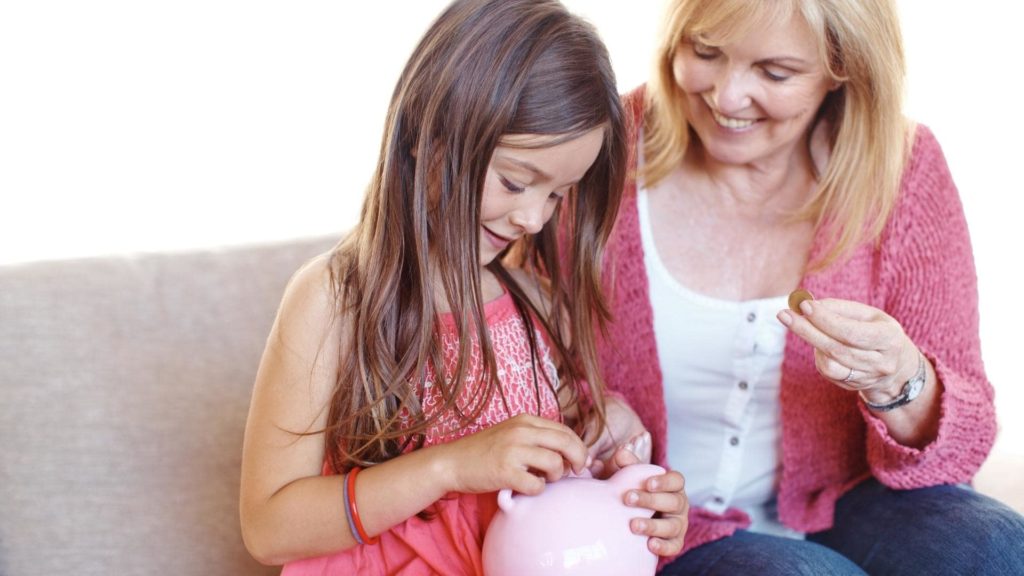 A young girl in a pink dress is placing a coin into a pink piggy bank, while an older woman smiles and holds another coin. They are sitting on a couch, engaged in a playful and educational activity.