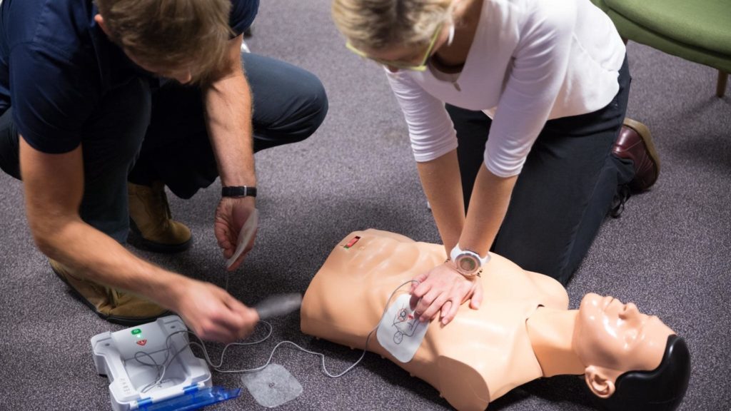 Two people are kneeling on the floor practicing CPR on a training mannequin. One person is performing chest compressions, while the other is using an AED. The mannequin is placed on a gray carpeted surface.