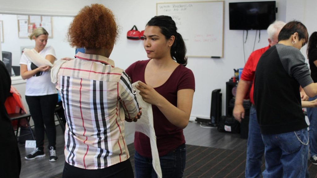 A group of people in a room practicing first aid techniques. One person is helping another wrap a bandage around their arm. Several others are engaged in similar activities in the background. A whiteboard and TV are mounted on the wall.
