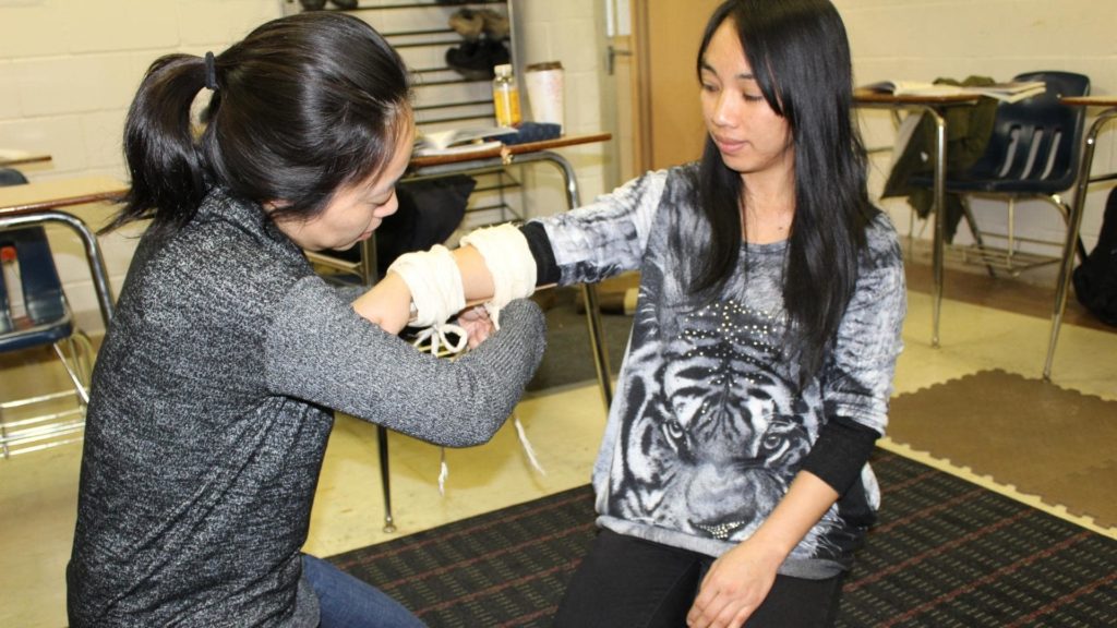 Two women are in a classroom. One is demonstrating a technique on the other's arm using a white wrap. They are seated on chairs, and desks are visible in the background. The woman on the right is wearing a tiger-print shirt.